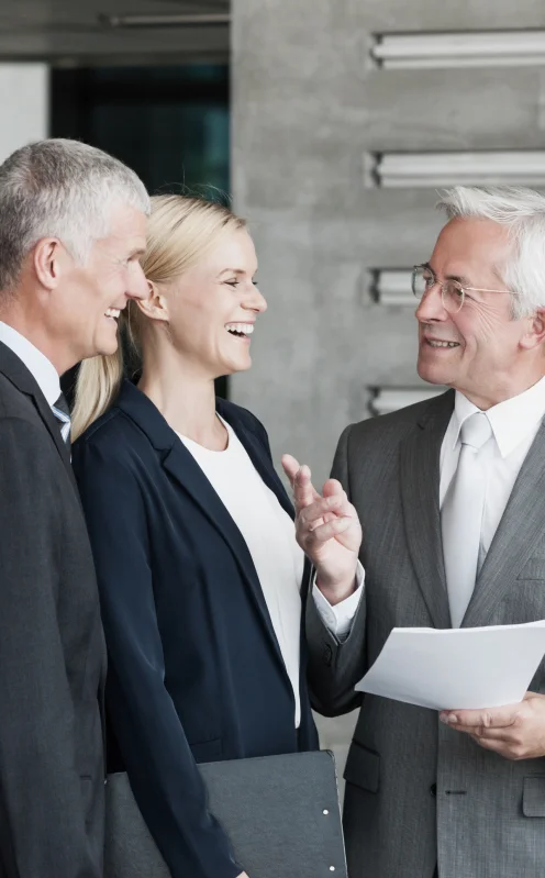 Three professionals engage in a discussion, with one gesturing and holding documents, set in a modern office environment.